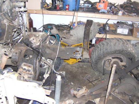 1989 Ford Bronco - A disassembled vehicle chassis with a large tire and engine parts is seen in a cluttered workshop environment.
