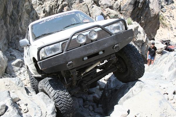 White 1993 Toyota 4Runner navigating rocky terrain with a custom bumper and large tires. Another vehicle is visible in the background