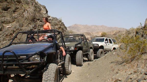 Several off-road vehicles, including a blue buggy and a white SUV, driving on a rocky trail in a desert landscape