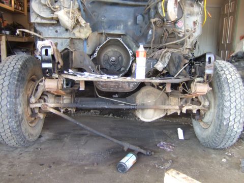 1989 Ford Bronco - The front chassis and drivetrain of a disassembled vehicle are visible, with the transmission housing exposed and suspension components lai…