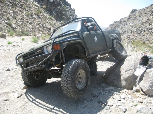 Modified Toyota pickup truck with large tires climbing over rocks on a trail. Has a roll cage and military-style star decal
