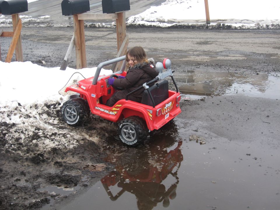 Red toy Jeep Wrangler with a child driving, parked in mud and puddles