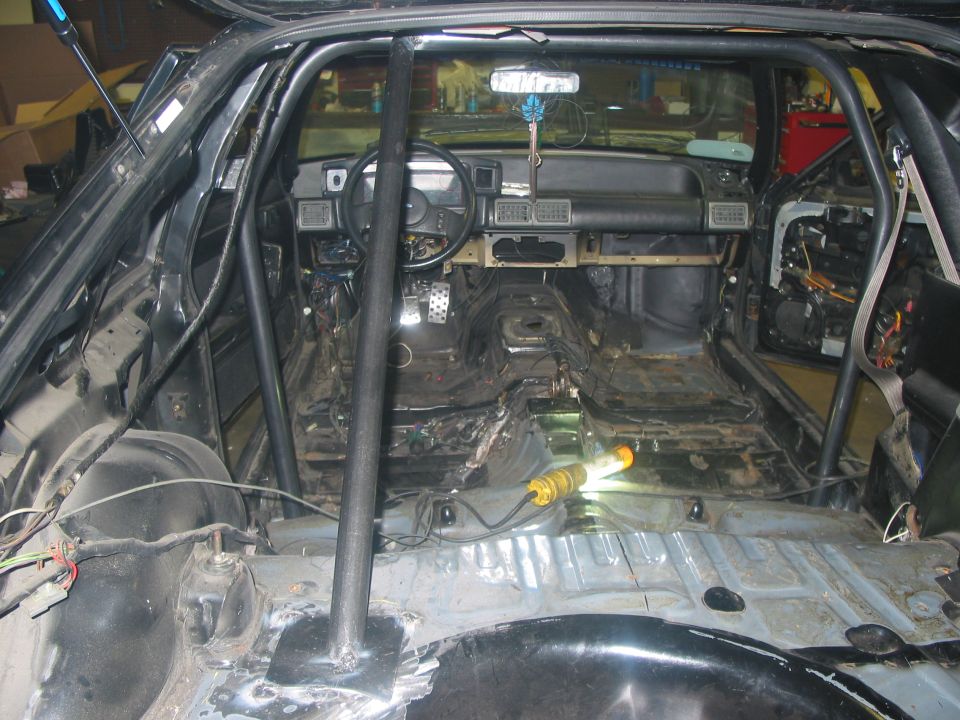 Gutted black Mustang interior with roll cage, bare floor pan, exposed wiring, and work light in a garage.