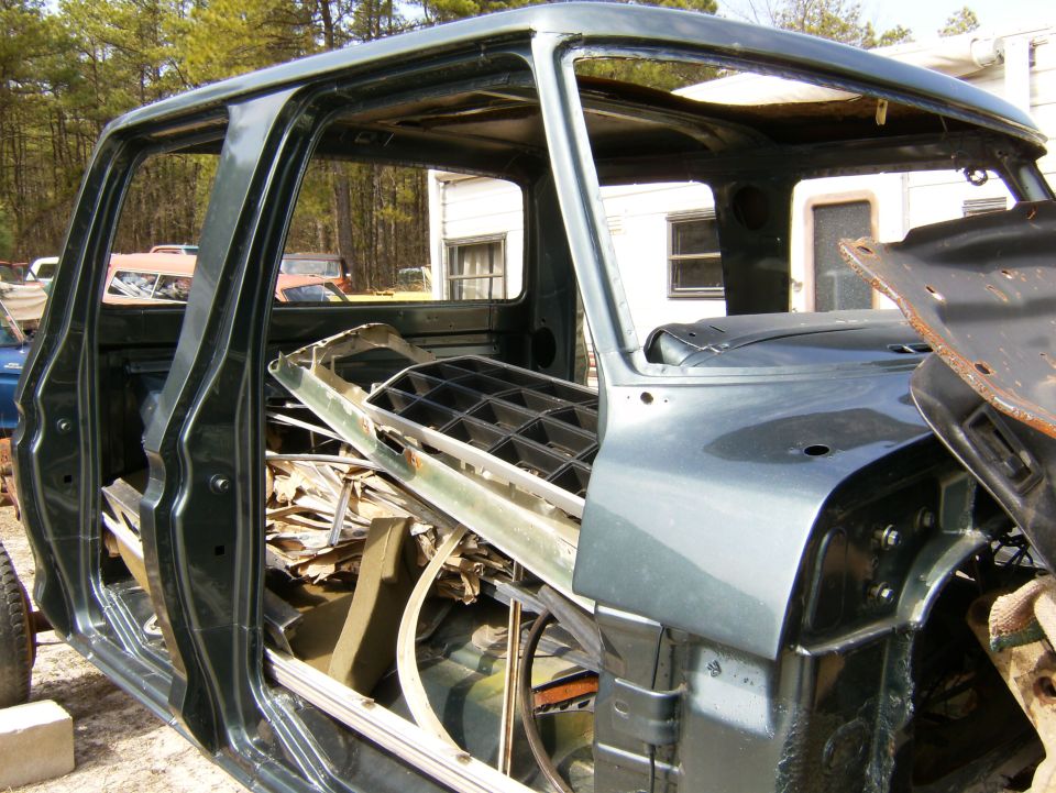 Bare dark green Ford crew cab body shell with parts piled inside, in a junkyard setting