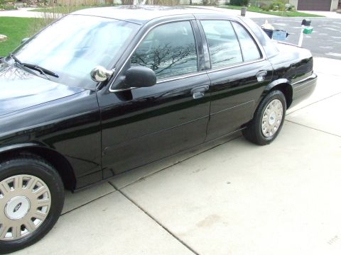 2003 Ford Crown Victoria - The side profile of a black 2003 Ford Crown Victoria sedan is shown parked on a concrete driveway. The car features chrome accents on the d…
