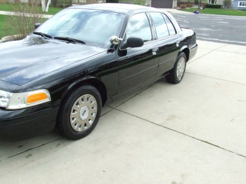 2003 Ford Crown Victoria - The driver's side of a black 2003 Ford Crown Victoria is shown parked on a concrete driveway. The car is a four-door sedan with chrome trim…