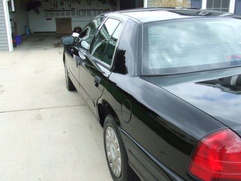 2003 Ford Crown Victoria - The rear passenger side of a shiny black sedan is shown parked on a concrete driveway in front of a garage. The car has chrome trim around…