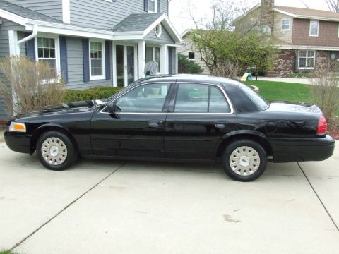 2003 Ford Crown Victoria - A black 2003 Ford Crown Victoria sedan is parked on a concrete driveway in front of a gray house with blue shutters. The car is viewed from…