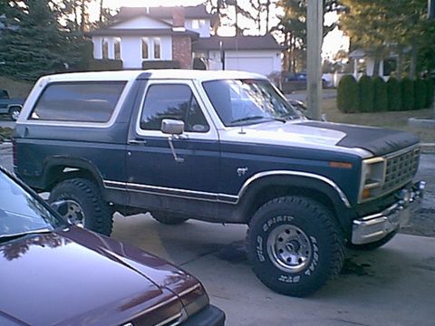 1981 Ford Bronco - A dark blue and white Ford Bronco is parked on a street in front of houses and trees.
