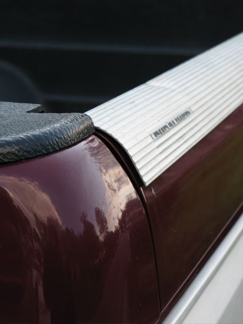 1991 Ford F-150 - A close-up view of the bed rail of a burgundy pickup truck, featuring a black textured bed rail cap and a silver ribbed bed rail cover with…