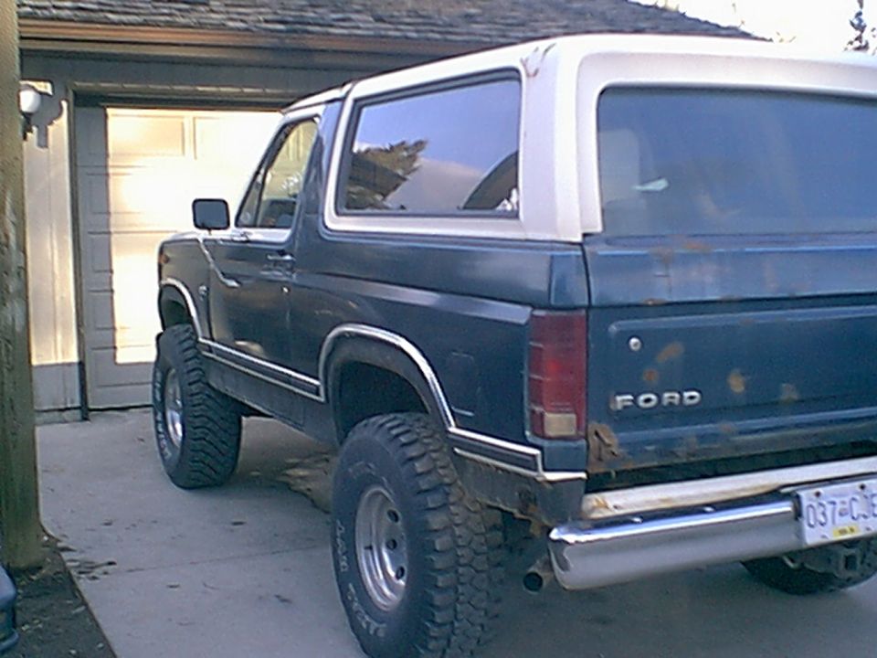 Dark blue 1980s Ford Bronco with white hardtop and rusty tailgate parked in a driveway