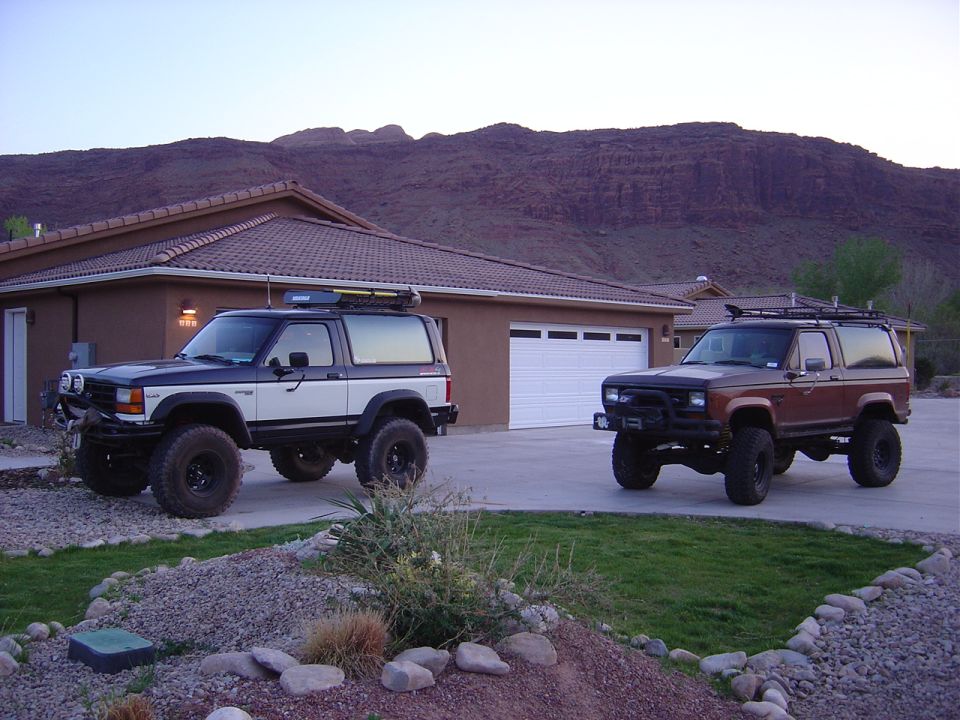 Two lifted Ford Bronco II SUVs parked in a driveway with a house and mountains in the background.