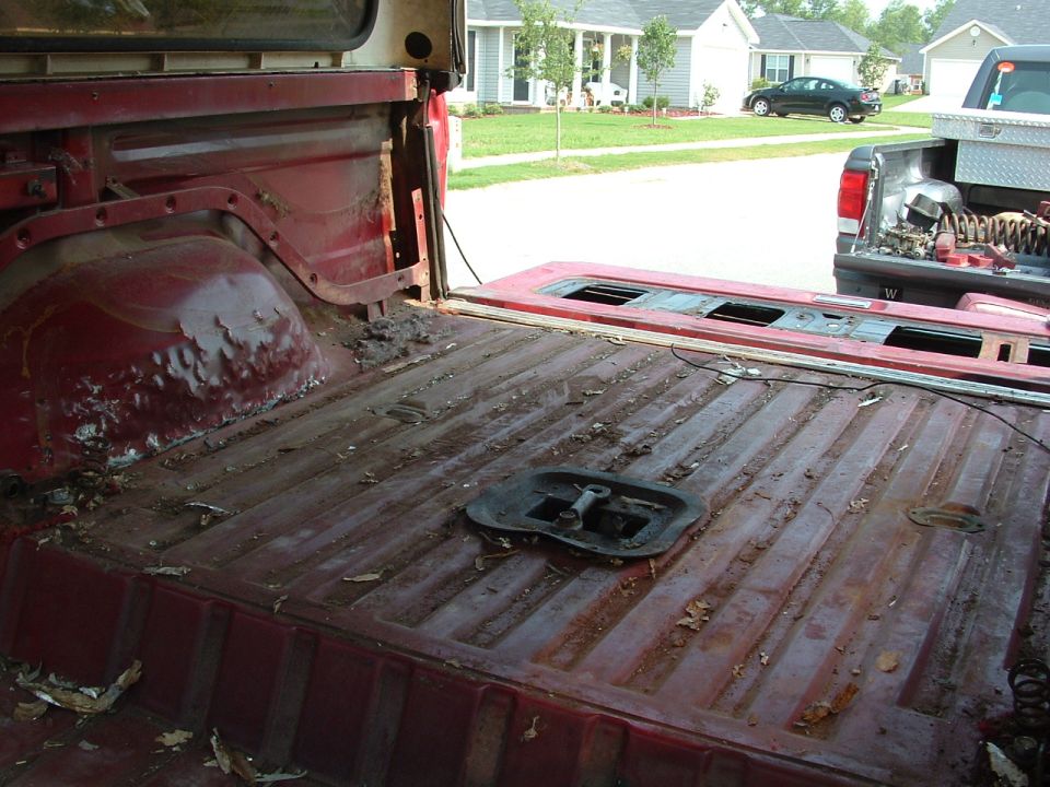 Interior view of a rusty red pickup truck bed with debris, another truck in background.