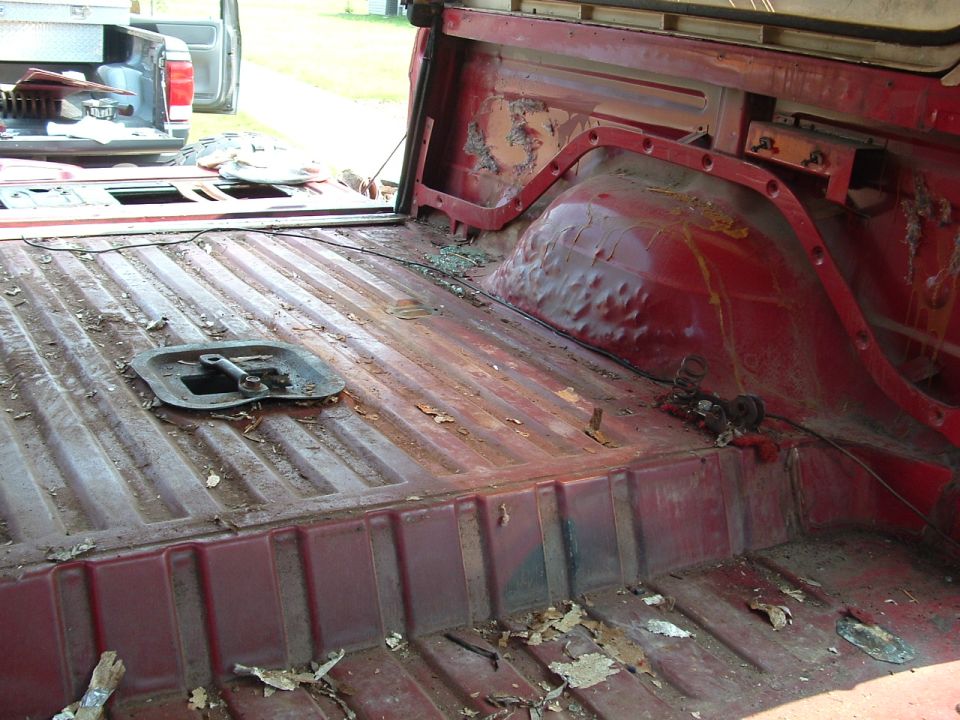 Interior of a red pickup truck bed with ribbed floor, rust, debris, and a metal bracket.