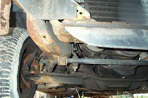 2001 Ford Crown Victoria - The underside of a vehicle reveals heavily corroded suspension parts and a visible tire, with the serpentine belt system in view.