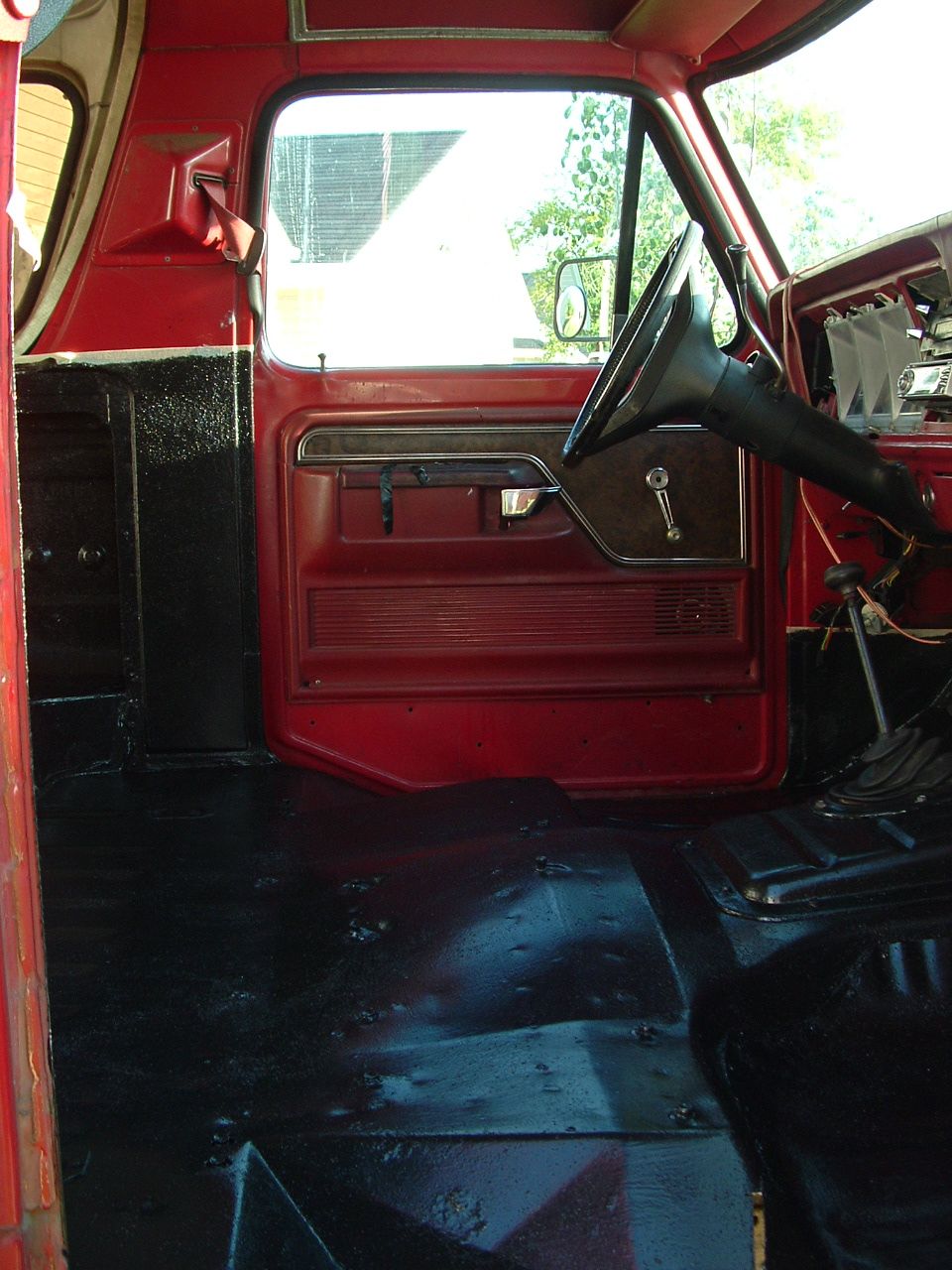 Interior view of a red classic Ford truck or SUV, showing the driver's door panel, steering wheel, and black floor.