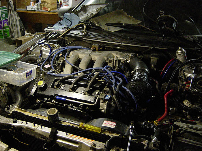 Transverse V6 engine mid-install with blue plug wires and cone air filter in a garage