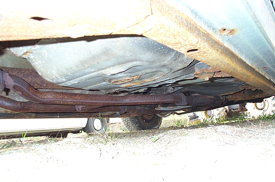 Underside view of a car, showing a rusty exhaust system and fuel tank. Some grass and dirt are visible below