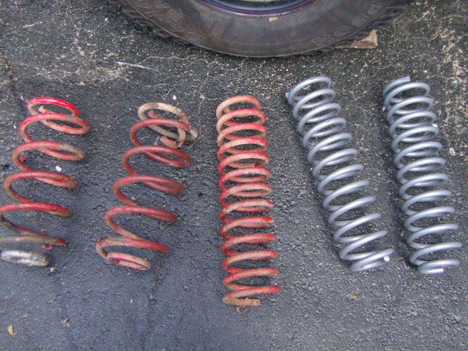 Five suspension coil springs, three red and two silver, laid out on asphalt.