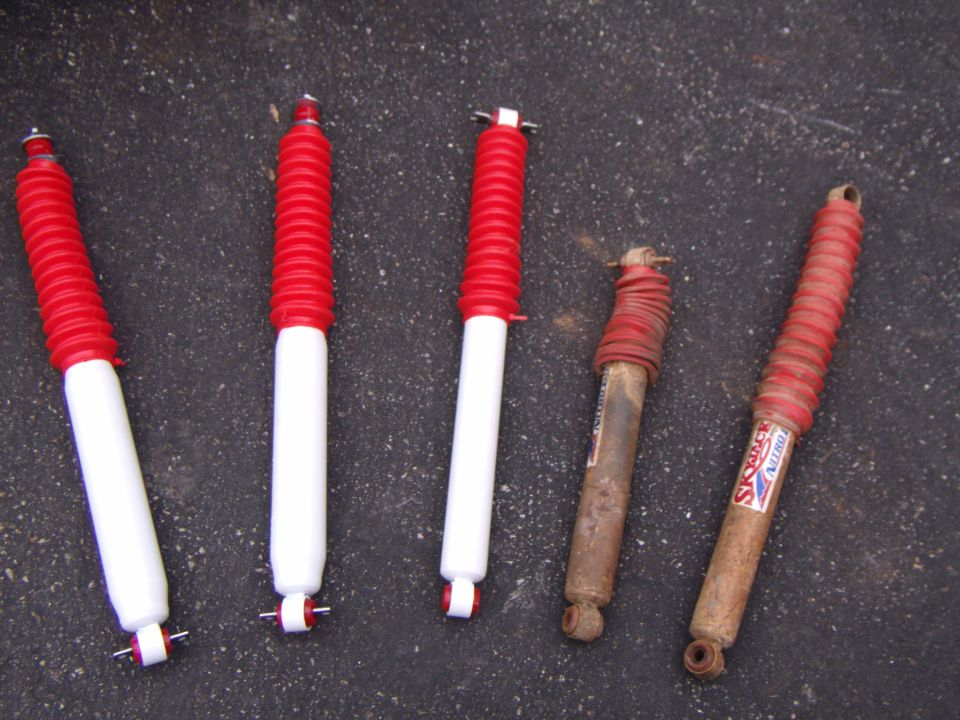 Three new white and red shocks beside two rusty muddy Skyjacker shocks on asphalt