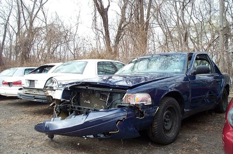 2001 Ford Crown Victoria - A damaged blue 2001 Ford Crown Victoria sits in a junkyard next to other vehicles.