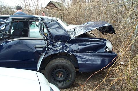 2001 Ford Crown Victoria - This heavily damaged blue sedan is partially obscured by brush and another vehicle.