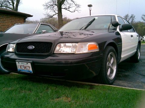 2003 Ford Crown Victoria - A black and white Ford Crown Victoria police car is parked on a wet asphalt driveway in front of a brick building and grass. The front of t…