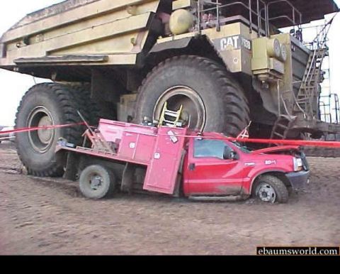 2000 Mercury Grand Marquis - A red service truck is dwarfed and stuck beneath the massive tires of a colossal yellow mining truck.