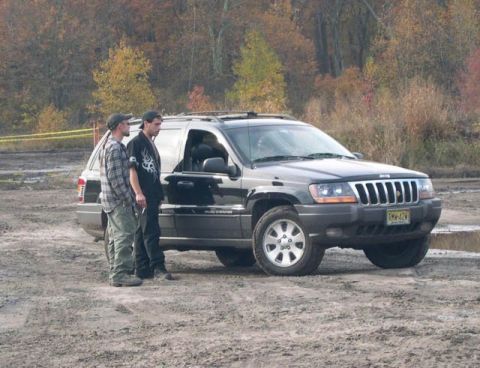 1990 Ford Bronco - Two men stand beside a black Jeep Grand Cherokee parked on a dirt lot with autumn foliage visible.