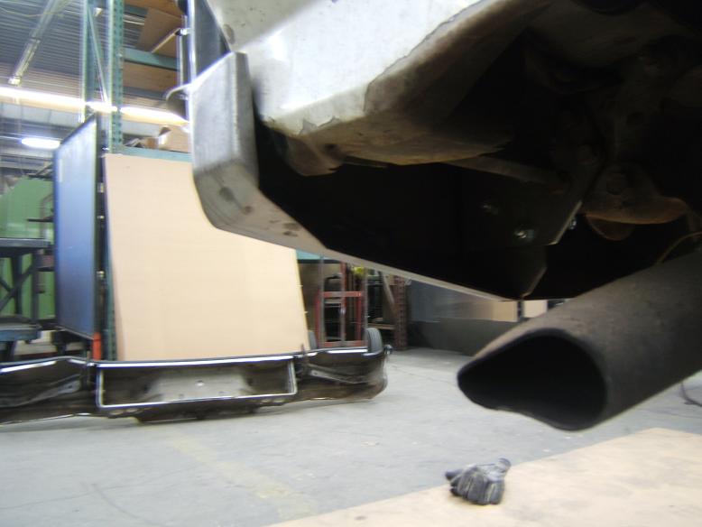 Underside view of a vehicle's rear bumper and exhaust pipe. A large cardboard panel is visible in the background