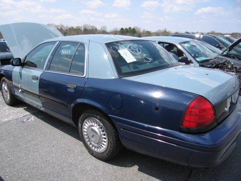2001 Ford Crown Victoria - A damaged 2001 Ford Crown Victoria sits in a junkyard with other vehicles.