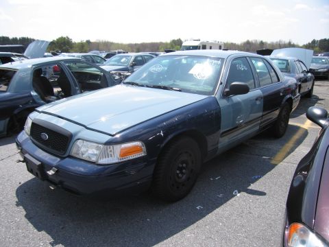 2001 Ford Crown Victoria - A two-tone blue Ford Crown Victoria is parked in a junkyard with its hood up, surrounded by other vehicles.