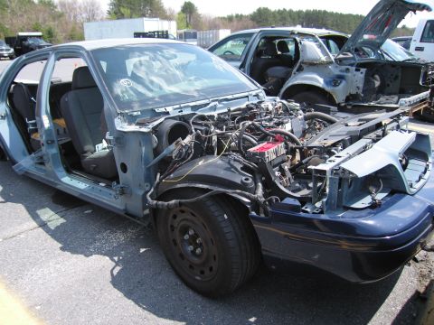 2001 Ford Crown Victoria - A dismantled 2001 Ford Crown Victoria sits in a junkyard with its hood open and engine exposed.
