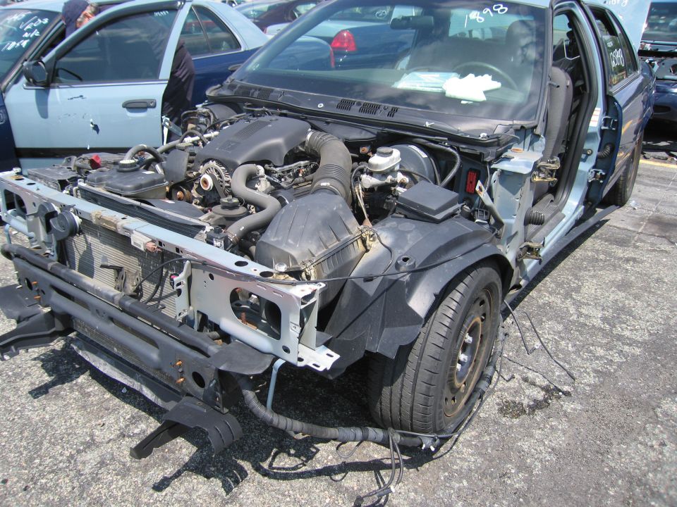 Front end of a 2001 Ford Crown Victoria with the hood open, showing the engine and missing body panels. Another car is visible to the left