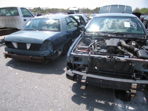 2001 Ford Crown Victoria - Two Ford Crown Victorias are parked in a lot, one with its hood open revealing the engine. Both vehicles appear to be damaged.