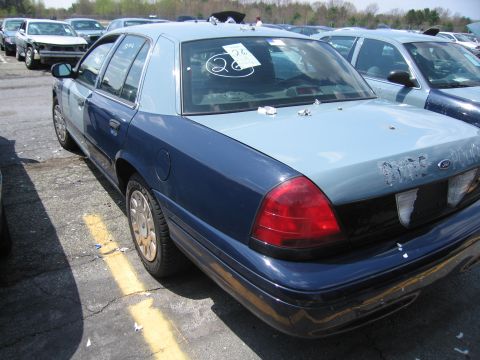 2001 Ford Crown Victoria - A two-tone police car is parked in a lot with other vehicles, showing a "26" sticker on the rear window.