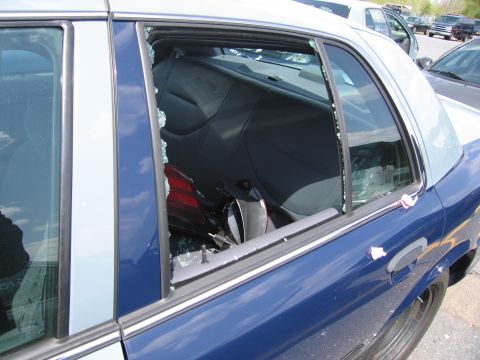 2001 Ford Crown Victoria - The rear passenger window of a police car has been broken, with glass fragments scattered inside and outside the vehicle.