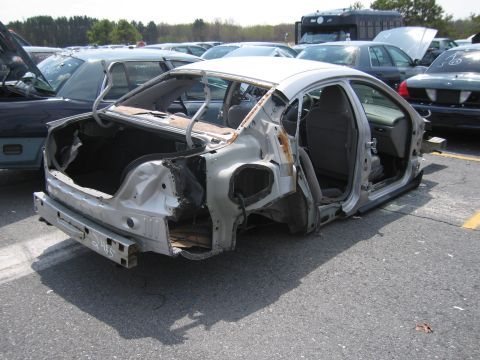2001 Ford Crown Victoria - A heavily damaged silver sedan sits in a junkyard, missing its trunk, doors, and most of its exterior.