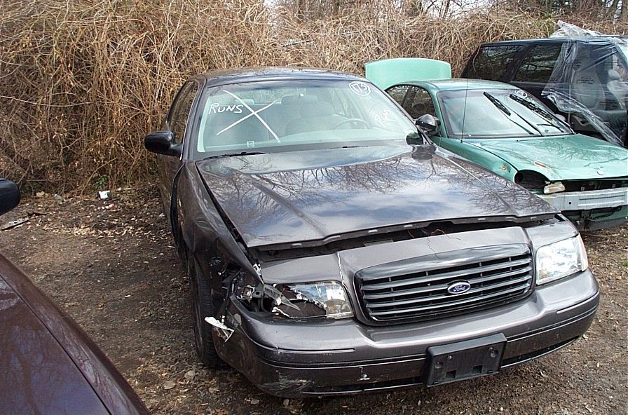 Front view of a dark gray 2001 Ford Crown Victoria with a damaged headlight and hood, parked next to a green car