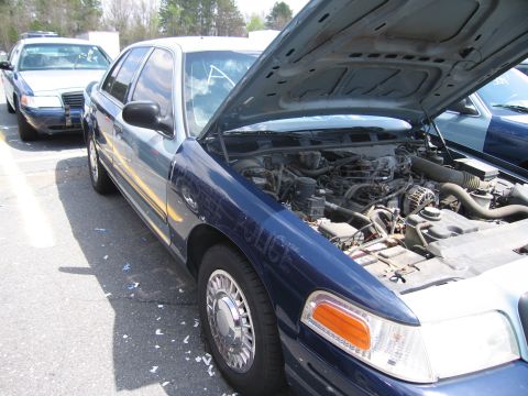 2001 Ford Crown Victoria - A blue police car with its hood open, showing the engine compartment. Another police car is visible in the background.