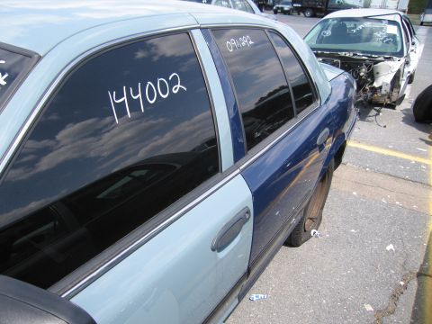 2001 Ford Crown Victoria - A two-tone sedan sits in a lot with a flat tire, displaying handwritten numbers on its windows.