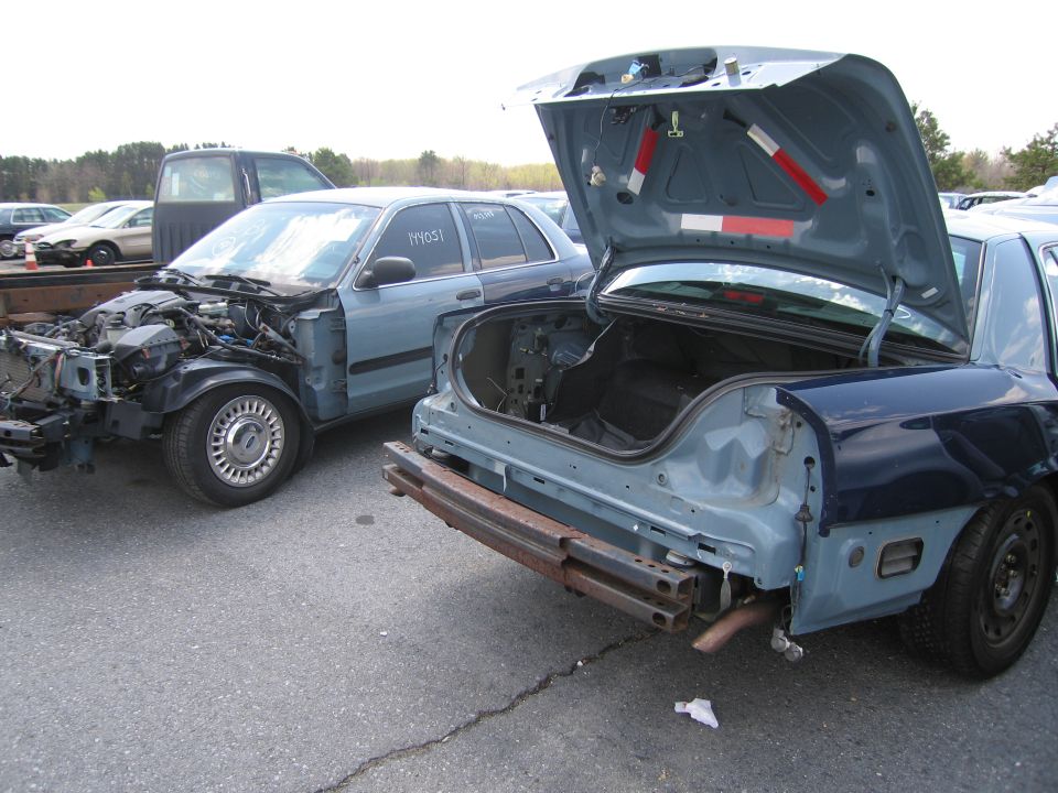 Two blue Ford Crown Victorias, one with its trunk open and rear bumper removed, the other missing its front end. Text '1Y105T' visible on the side of the second car