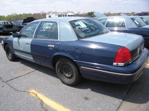 2001 Ford Crown Victoria - This two-tone police car, likely a 2001 Ford Crown Victoria, is seen from the rear quarter angle with visible markings on the window.