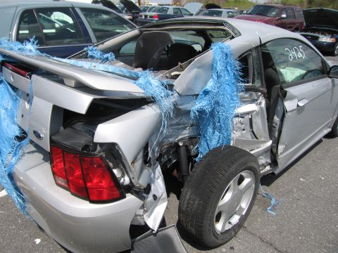 2001 Ford Crown Victoria - A severely damaged silver coupe sits in a junkyard with blue tarp remnants and a detached tire.