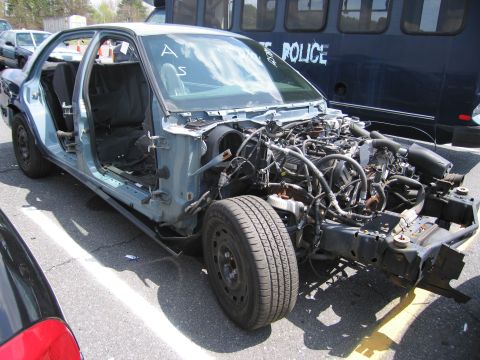 2001 Ford Crown Victoria - This stripped-down sedan shows its exposed engine and interior, with a police bus visible in the background.