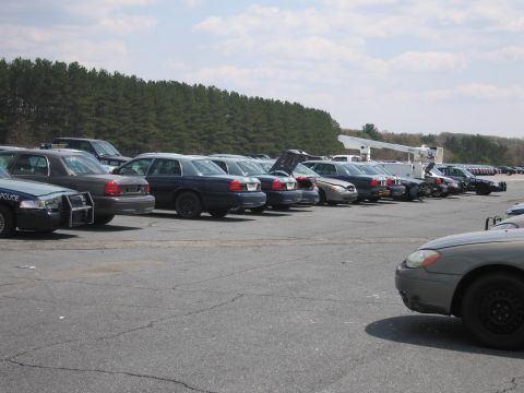 2001 Ford Crown Victoria - A collection of police vehicles and other cars are parked in an outdoor lot under a partly cloudy sky.