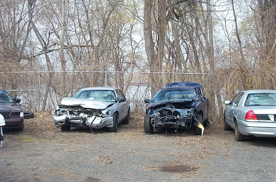 Two wrecked Ford Crown Victoria police cars parked side-by-side, one silver and one dark blue. The silver car is on the left, the dark blue car is on the right. Both have heavily damaged front ends