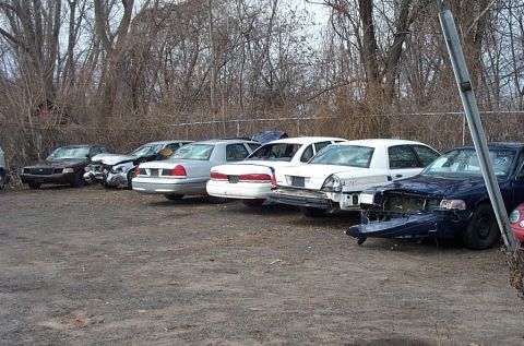 2001 Ford Crown Victoria - A line of damaged Ford Crown Victoria police cars are parked in a lot with trees in the background.