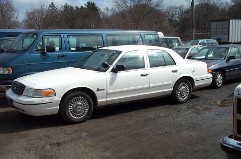 2001 Ford Crown Victoria - A white 2001 Ford Crown Victoria sedan is parked next to a blue van and other vehicles.
