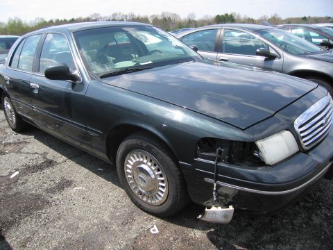 2001 Ford Crown Victoria - A dark green 2001 Ford Crown Victoria sits in a lot with other vehicles, showing damage to its front end.
